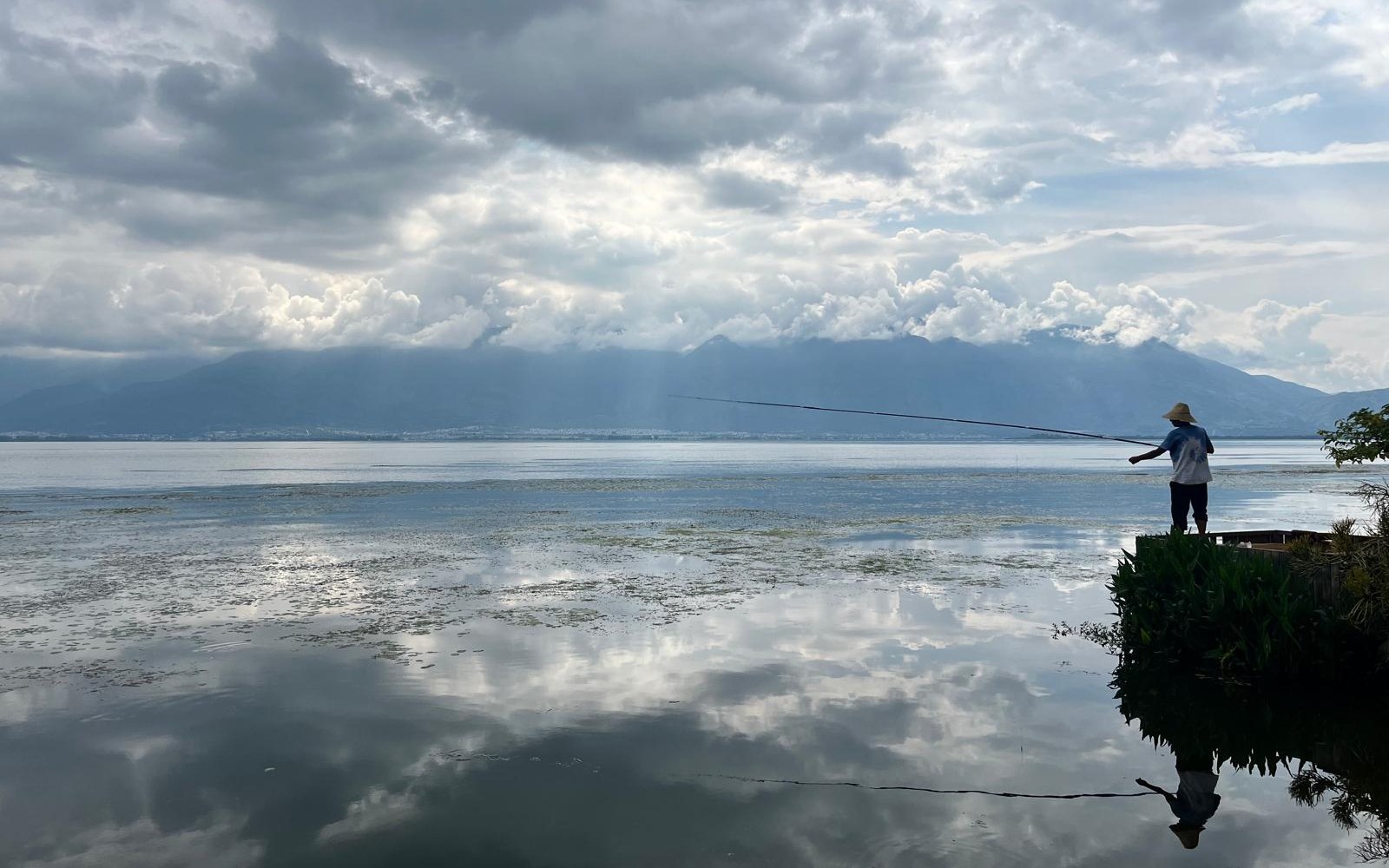 A fisherman beside a calm lake beneath a cloudy sky.